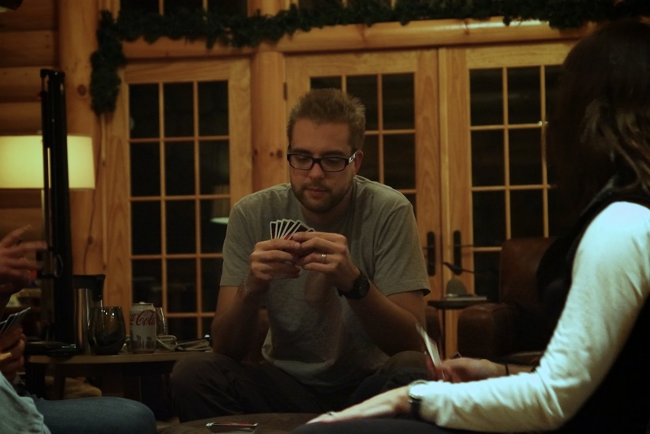 A man wearing glasses and a gray shirt focuses on his cards while playing a game with others.