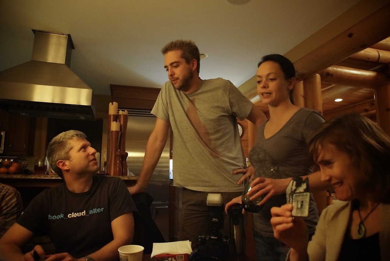 A group of people in a cabin-style kitchen, engaged in conversation and holding drinks and a card.