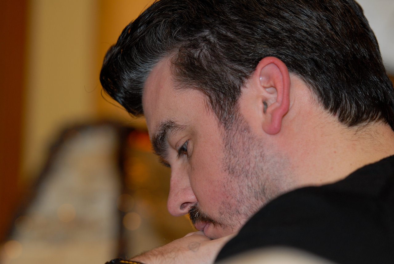 A man with dark hair and a beard is resting his head on his arm, looking downward.