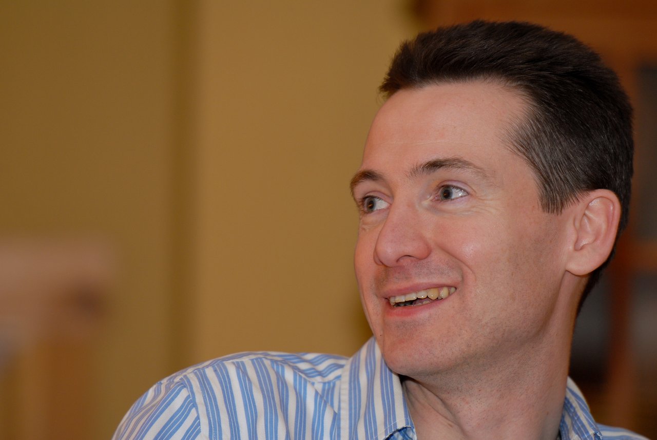 A man in a striped shirt smiles and looks to the side in an indoor setting.