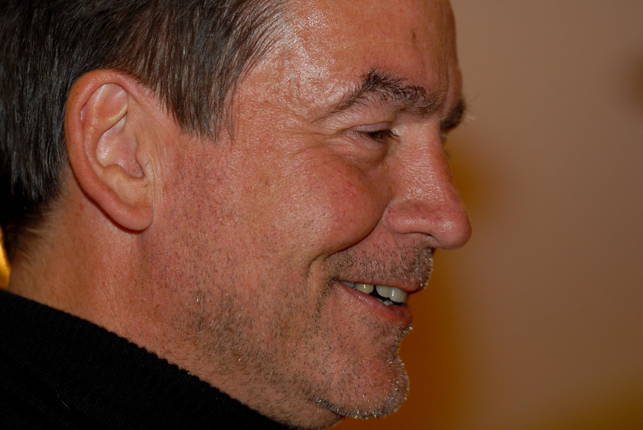 Close-up of a man in a black sweater smiling and looking to the side in a warm indoor setting.