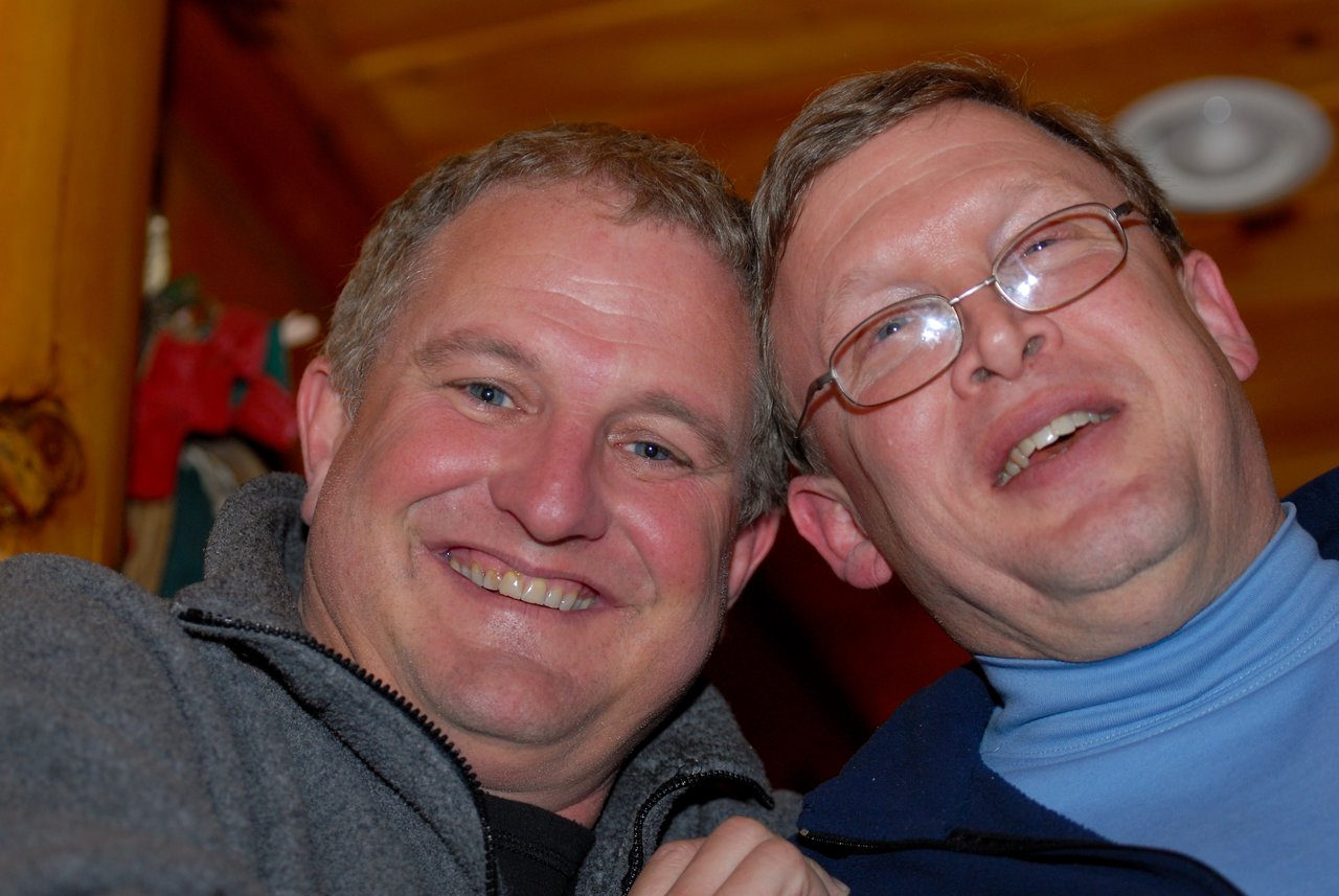 Two smiling men pose closely together indoors, looking at the camera.