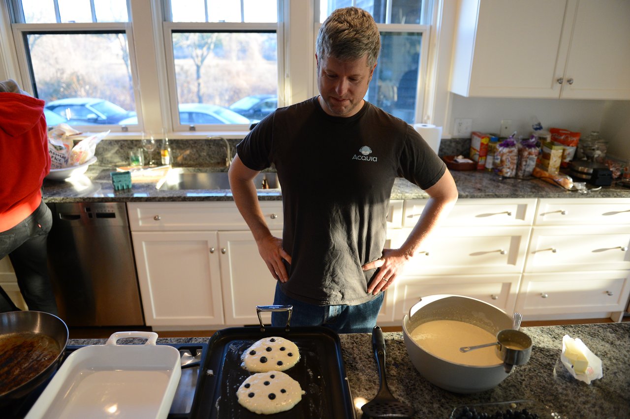 A man in an Acquia shirt looks at blueberry pancakes cooking on a griddle in a kitchen.