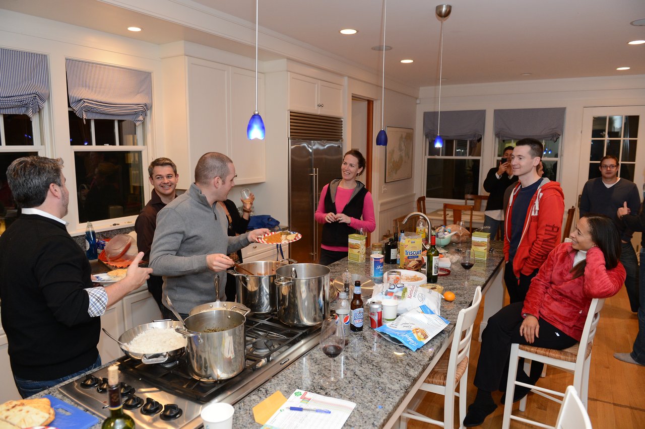 A group of people gathers in a kitchen, chatting and serving food from large pots on the stove.