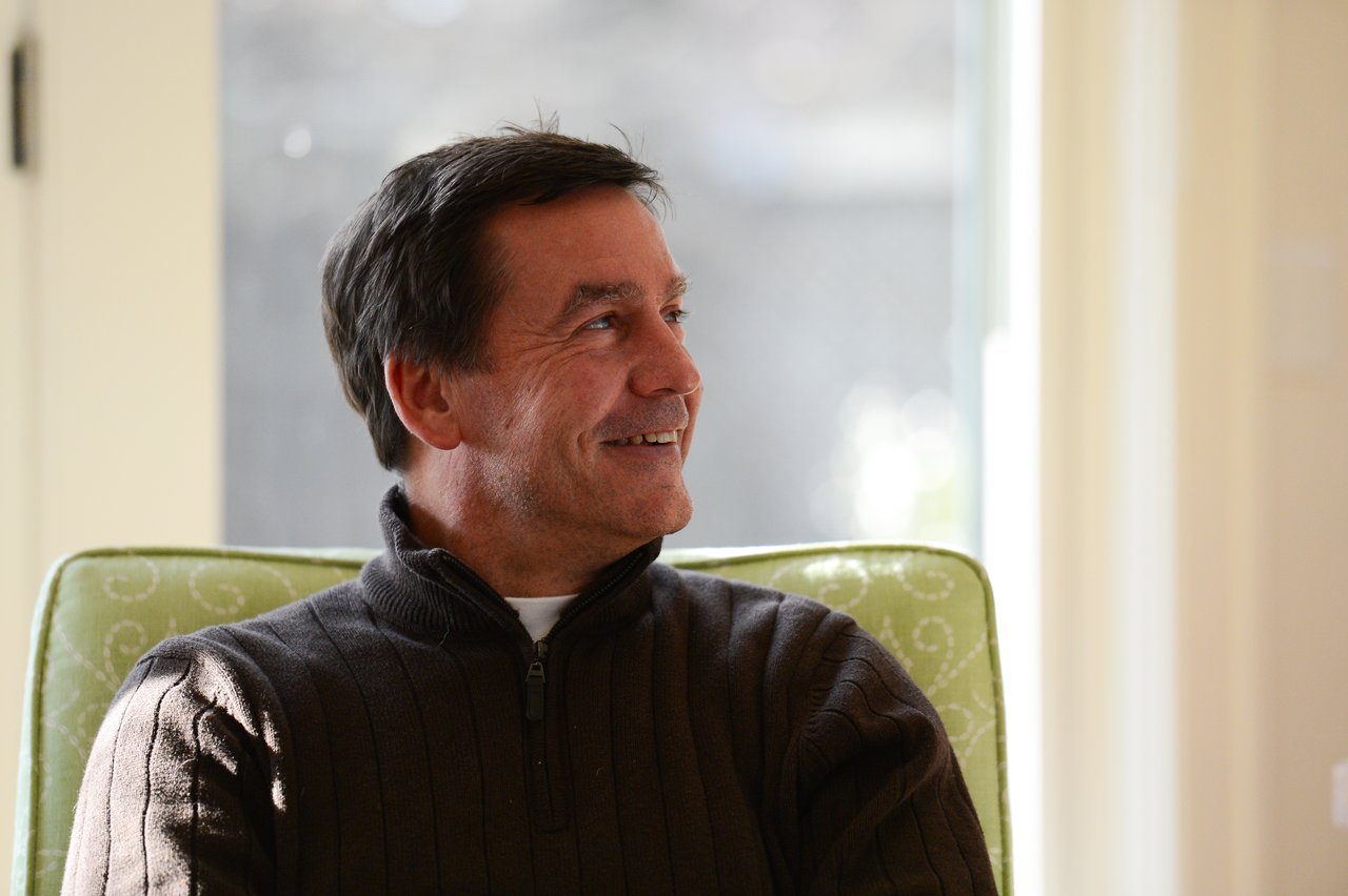 A man in a dark sweater sits on a green chair, smiling and looking to the side.