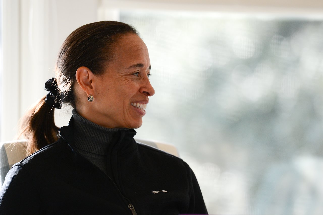 A woman in a black jacket sits indoors, smiling and looking to the side.