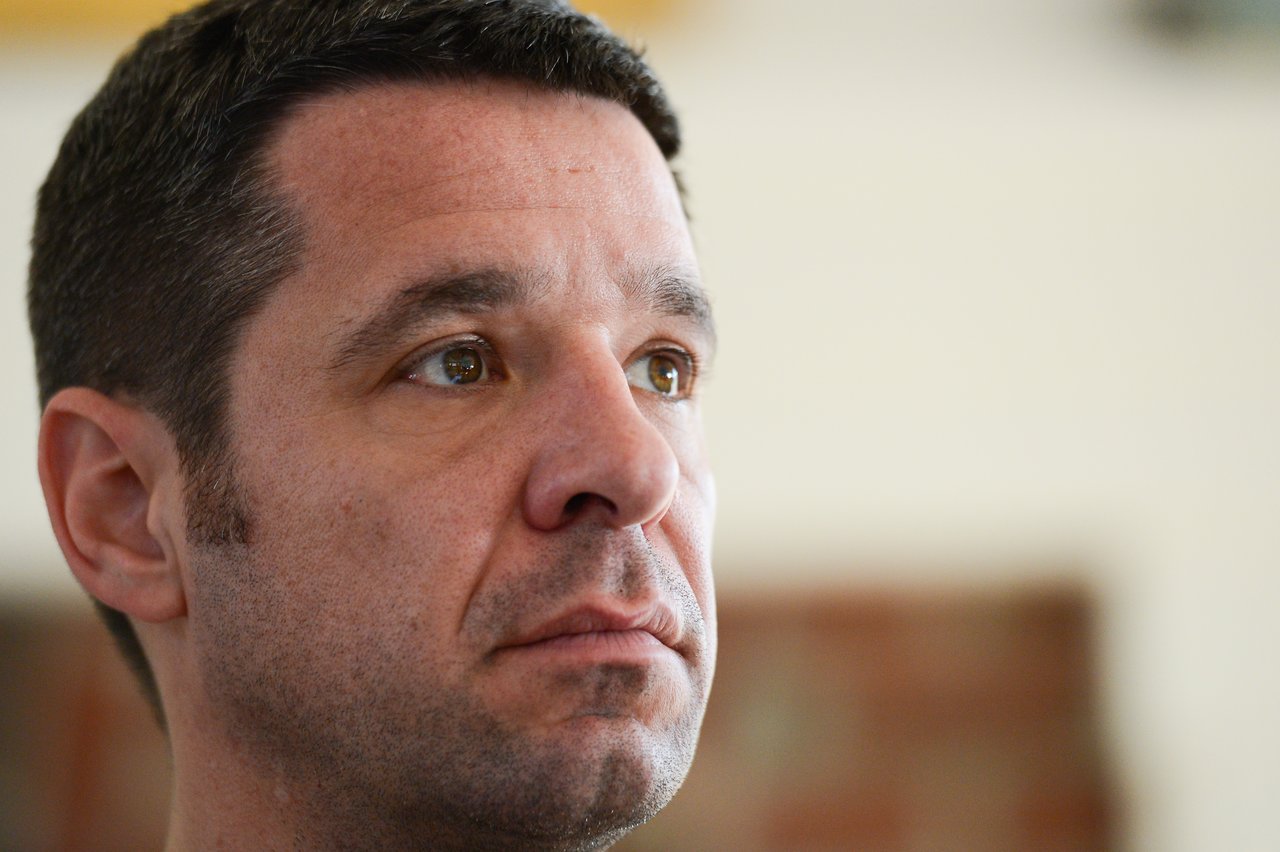 A man with short dark hair looks intently off-camera with a serious expression.