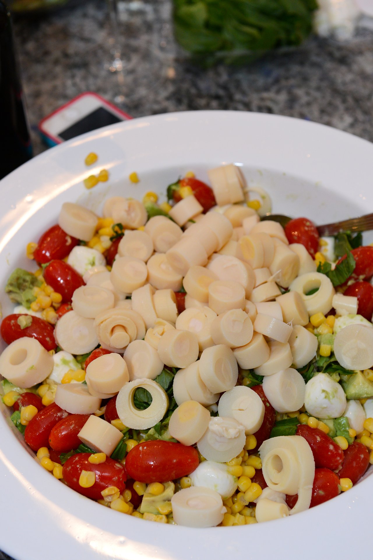 A white bowl filled with salad containing cherry tomatoes, corn, mozzarella, avocado, and sliced hearts of palm.