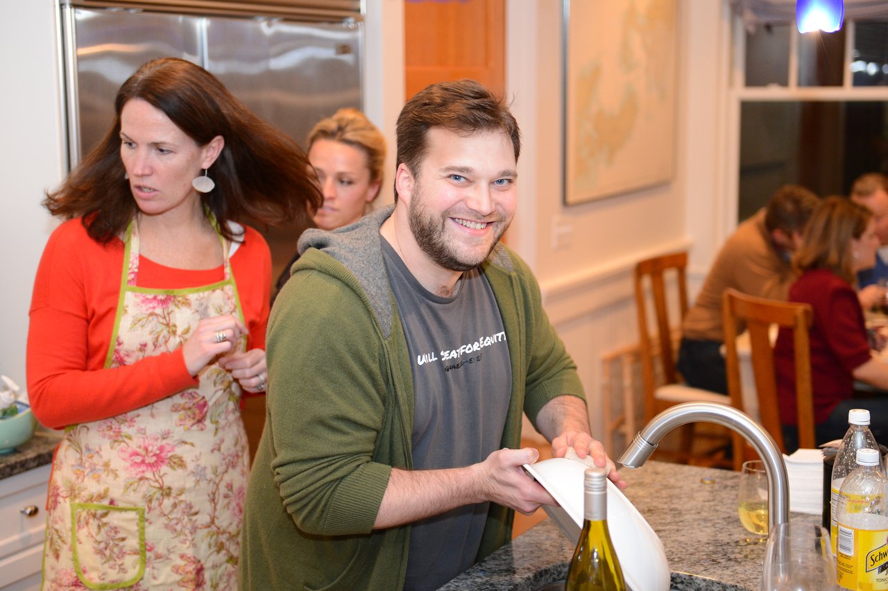 A smiling man washes dishes at a kitchen sink while others prepare food and talk in the background.