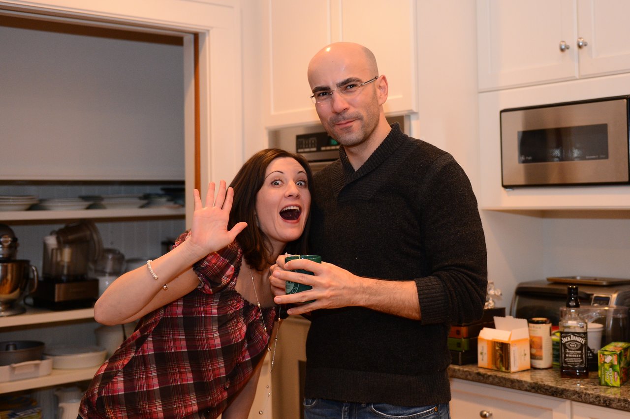 A woman playfully waves while a man holds a drink in a kitchen, both looking at the camera.