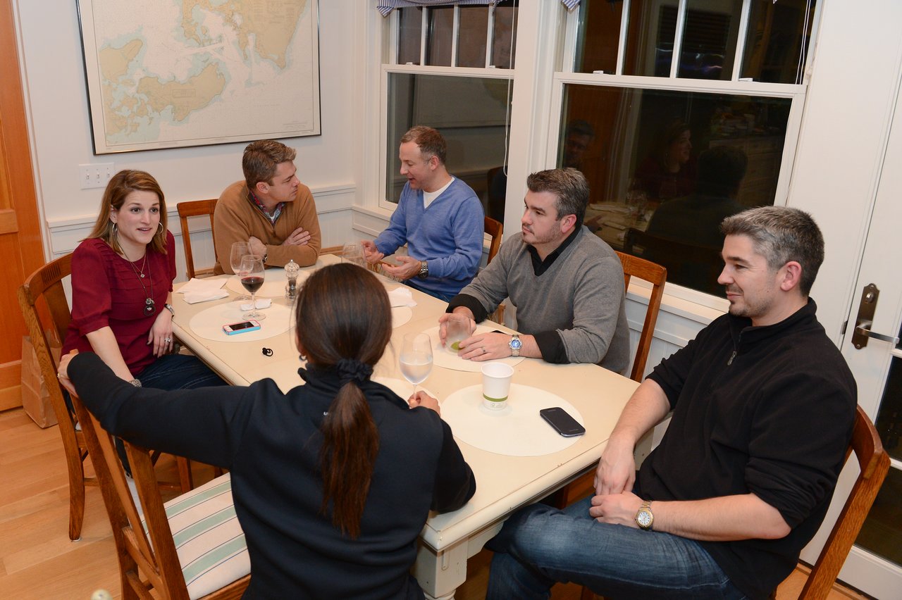 A group of six people sit around a table, engaged in conversation with drinks and phones nearby.