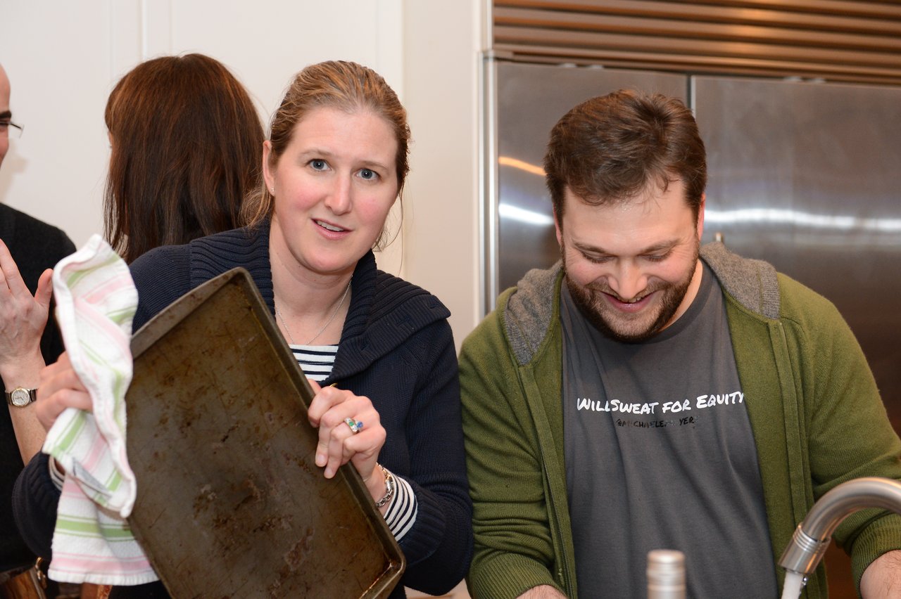 A woman holds a baking sheet while a man smiles and washes dishes in a kitchen.