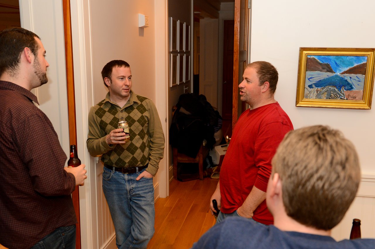 Four people stand in a hallway, holding drinks and engaged in conversation.