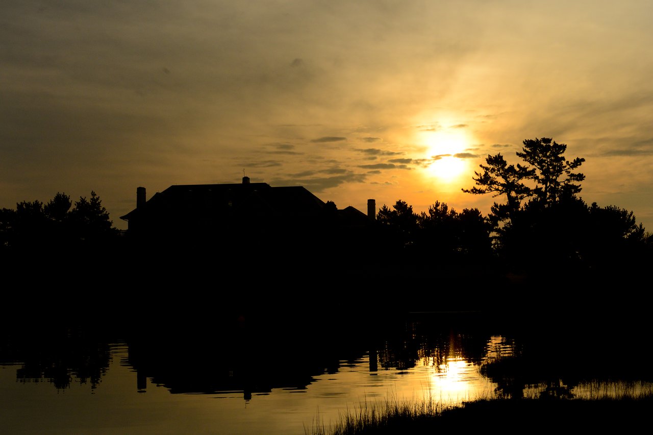 Silhouetted house and trees reflected in water at sunset, with a golden sky and scattered clouds.