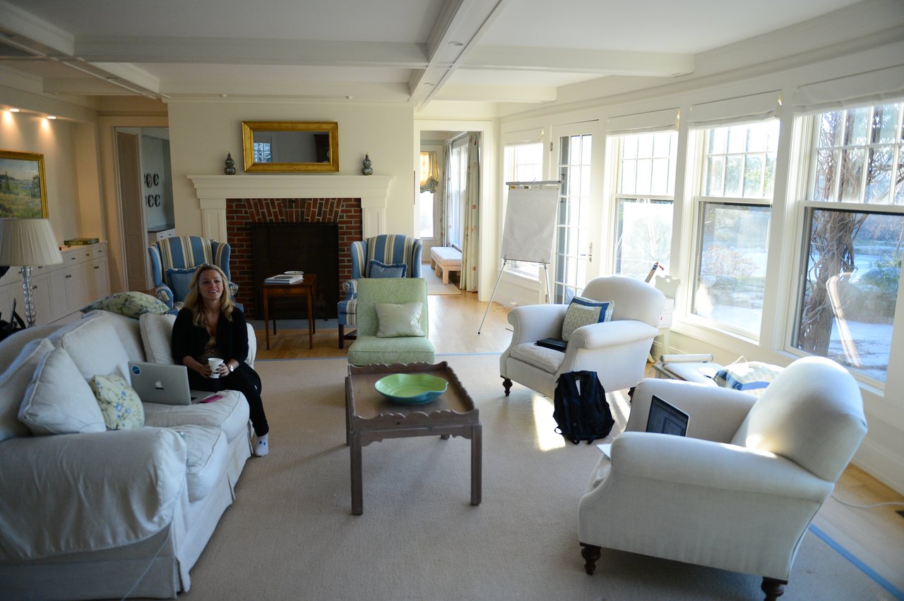 A woman sits on a couch with a laptop, holding a cup, in a bright living room.
