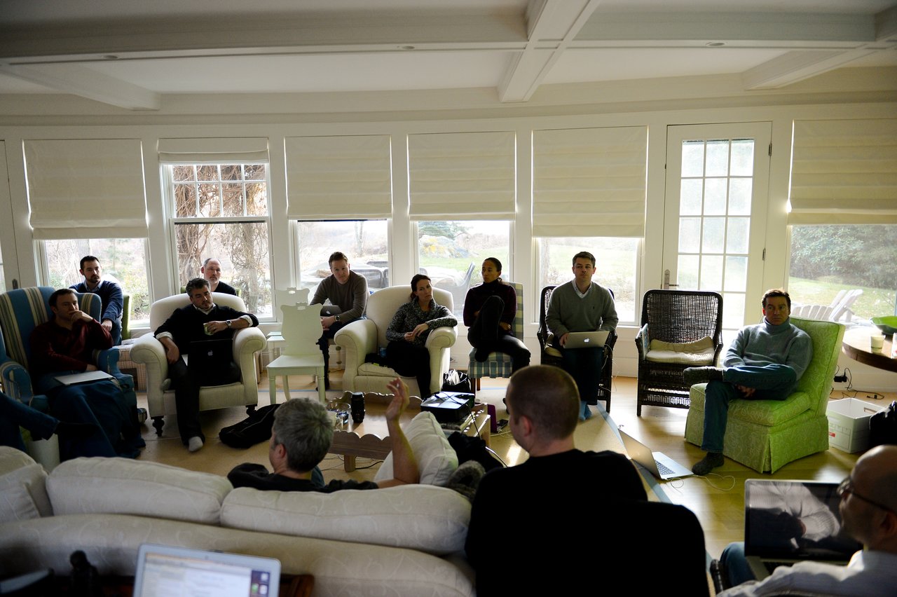 A group of people sit in a living room, listening to a speaker gesturing while leading a discussion.