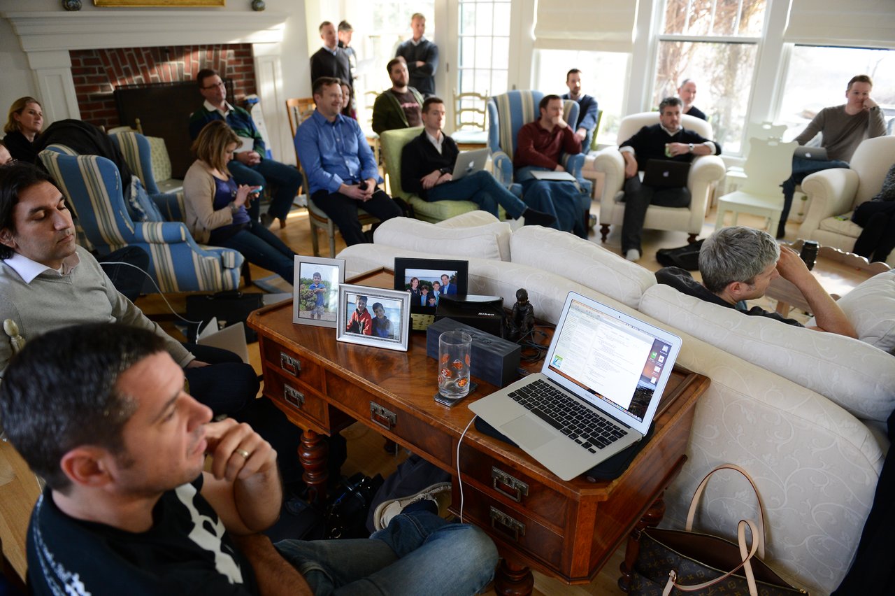 A group of people in a living room attentively listening to a presentation, with laptops and notebooks open.