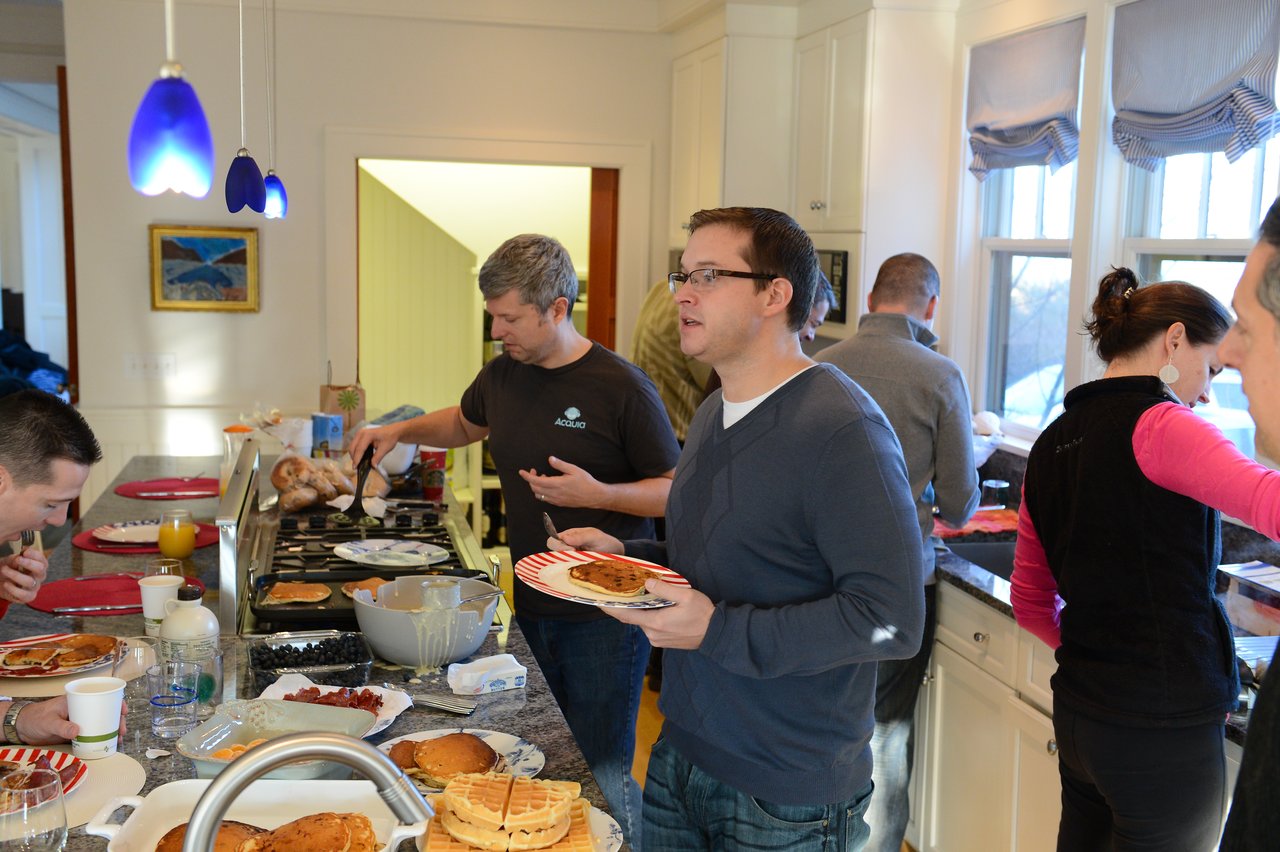 People gather in a kitchen, serving and eating pancakes.