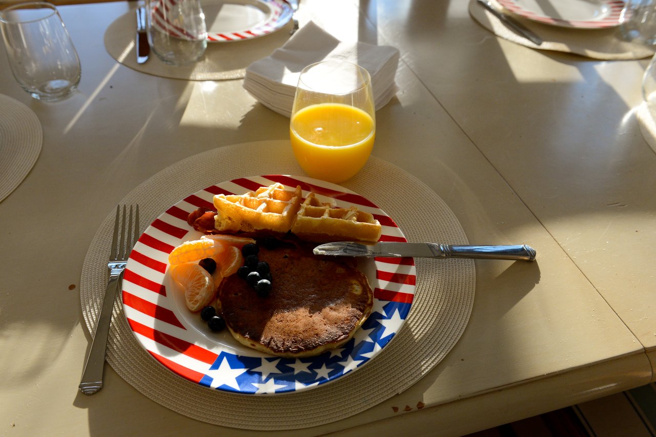 A breakfast plate with pancakes, waffles, bacon, fruit, and a glass of orange juice on a dining table.
