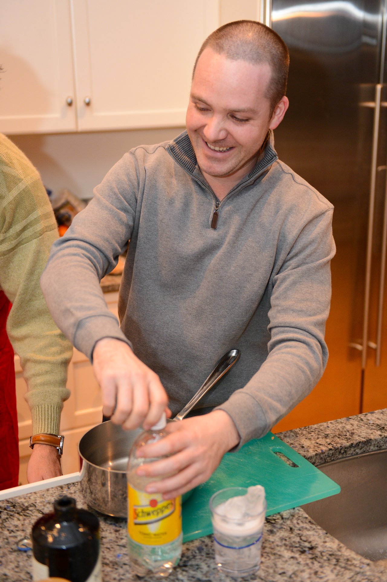 A smiling man in a gray sweater opens a bottle of Schweppes while preparing a drink in a kitchen.