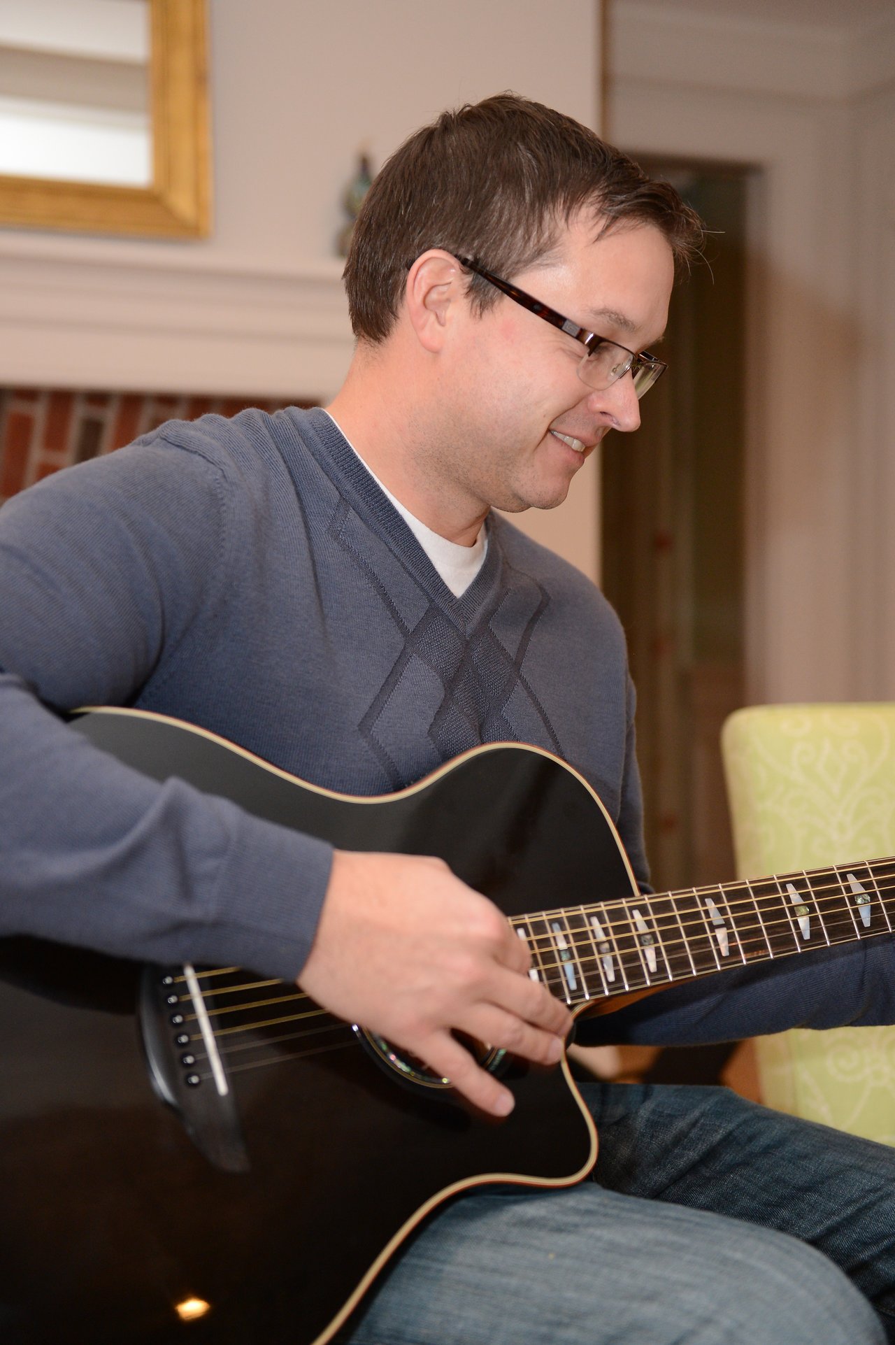 A person wearing glasses and a sweater plays a black acoustic guitar indoors, smiling while strumming.