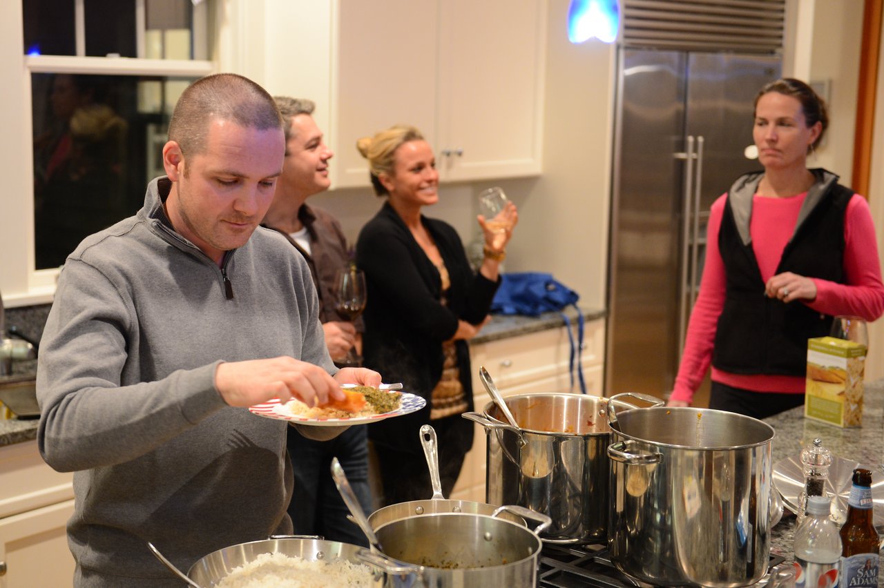 A man serves himself food from a pot while three people talk in a kitchen.