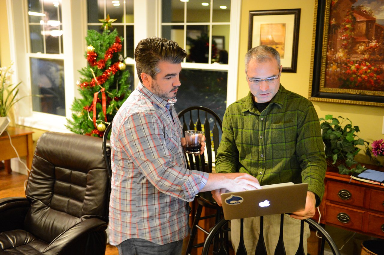 Two men stand near a laptop, one pointing at the screen while the other listens and holds a drink.