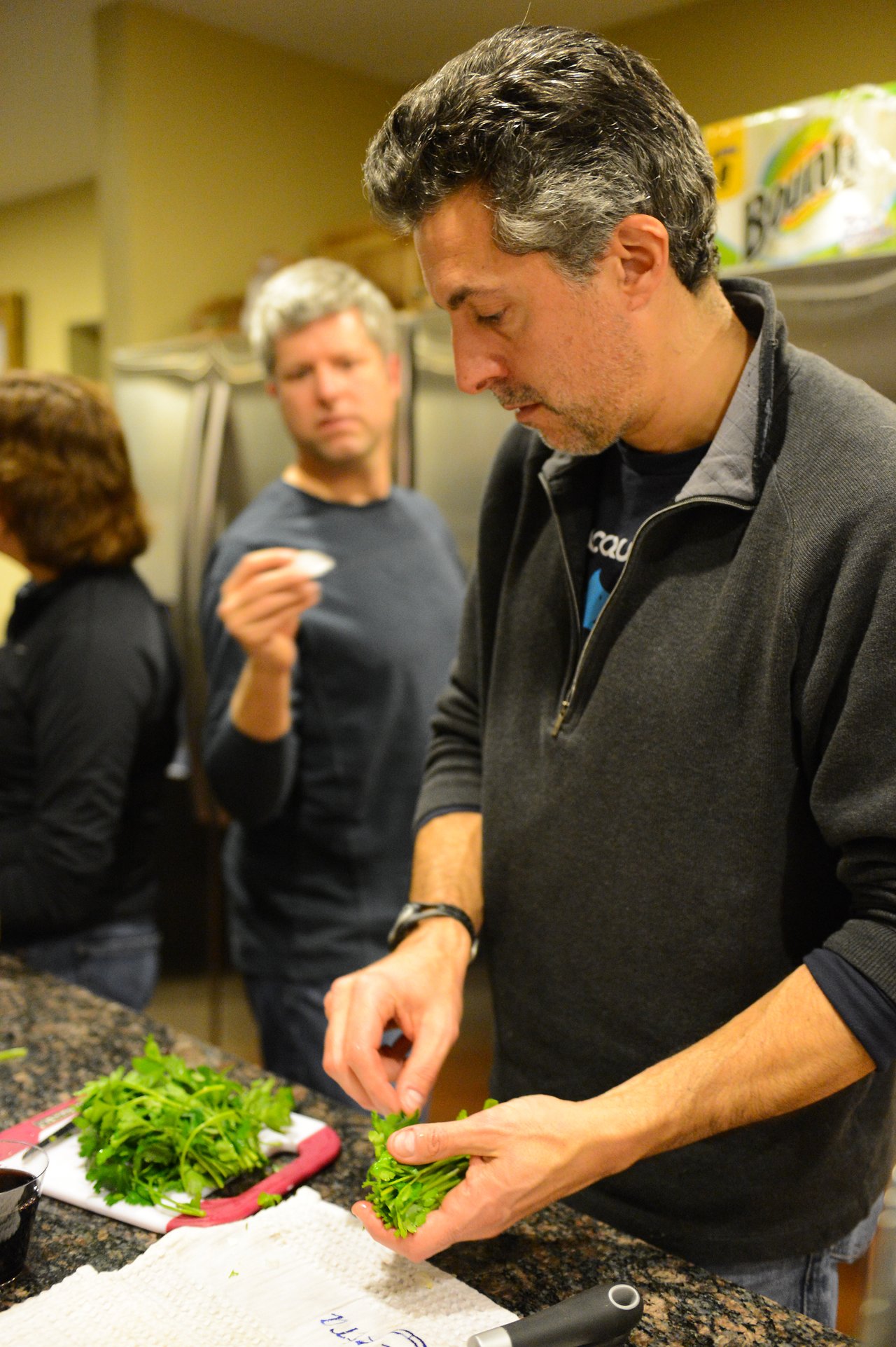 A man in a black jacket chops fresh herbs on a kitchen counter while others stand nearby.