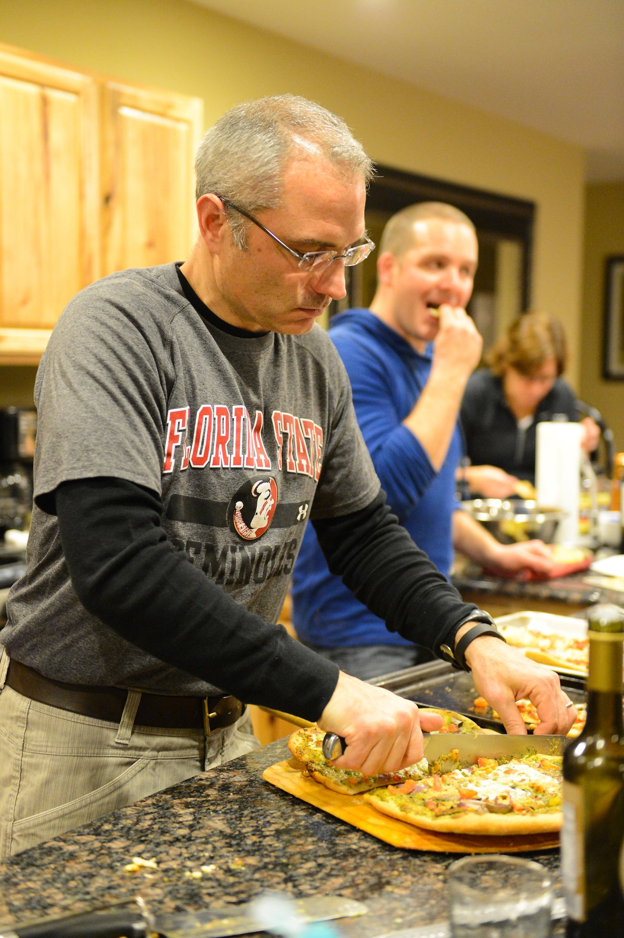 A man slices a freshly baked pizza on a cutting board while others eat and chat in the background.