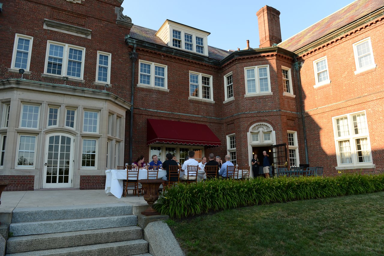 A group of people sits at a long outdoor table, having dinner near a large brick building.