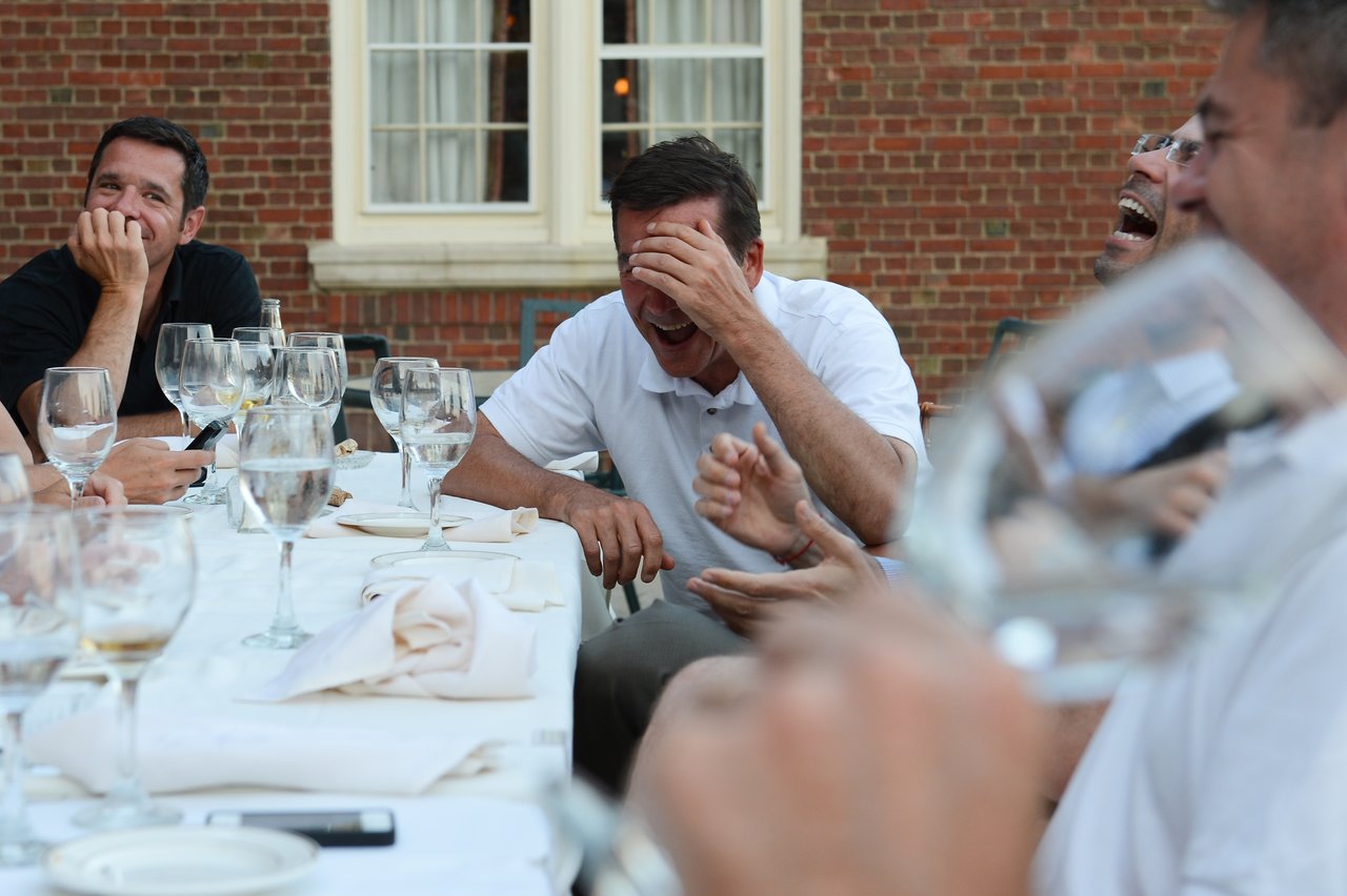 A group of people at an outdoor dinner table laughing, with one person covering their face while laughing.