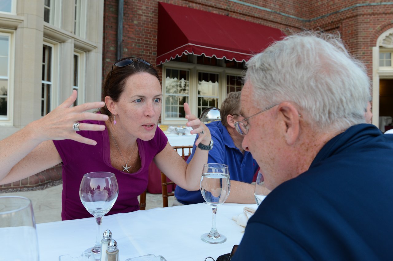 A woman in a purple shirt gestures expressively while speaking to an older man at an outdoor dinner table.