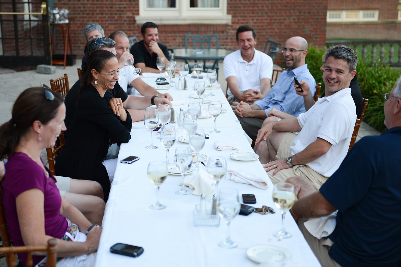 A group of people sit around a long table outdoors, smiling and laughing while enjoying drinks and conversation.