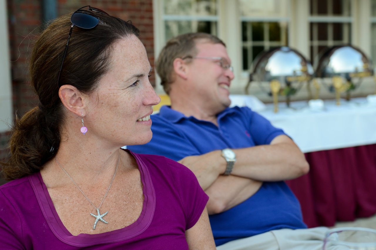 A woman in a purple shirt looks to the side while a man in a blue shirt sits smiling behind her.