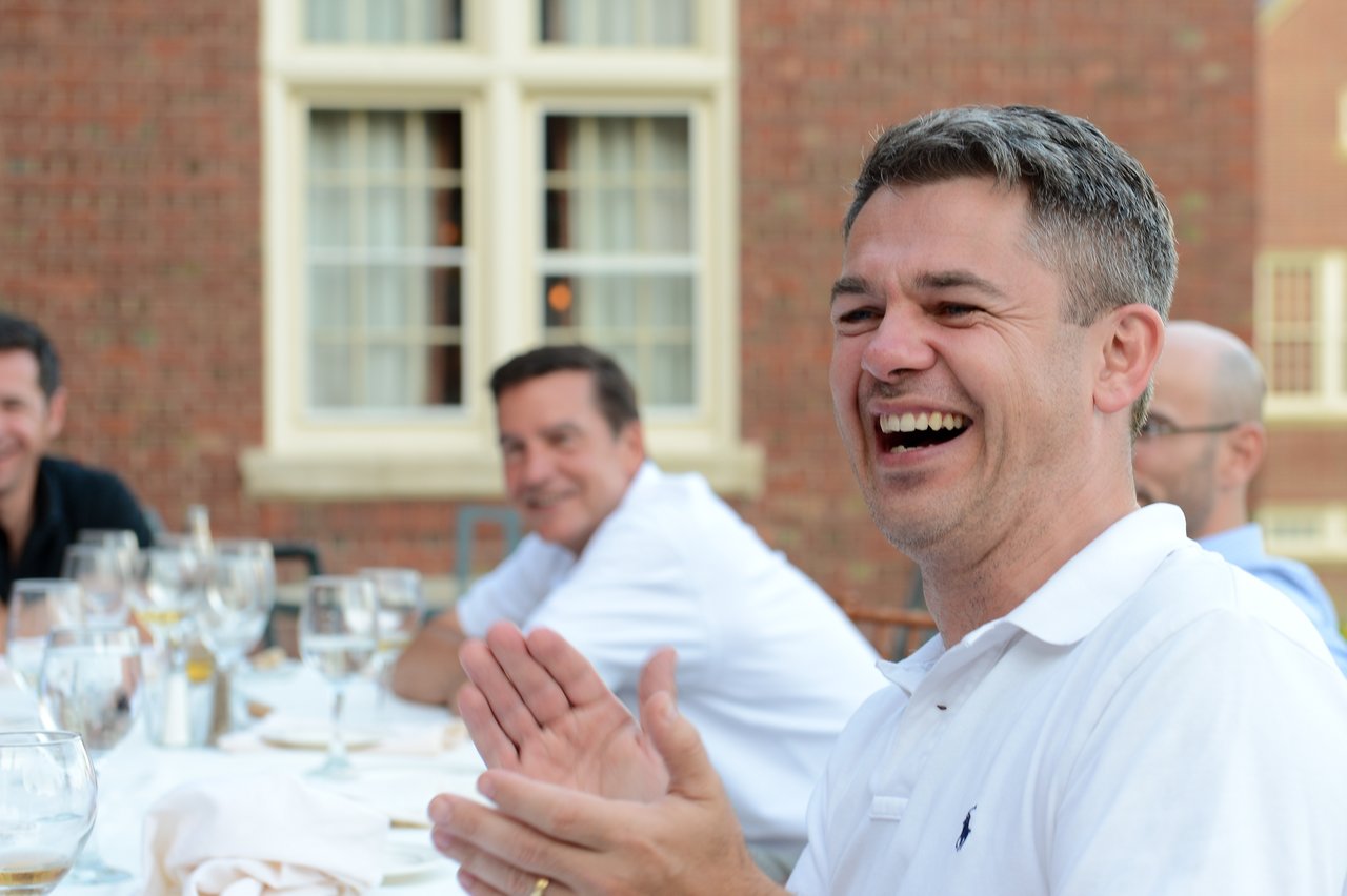 A man in a white polo shirt laughs and claps while seated at an outdoor dinner with others.