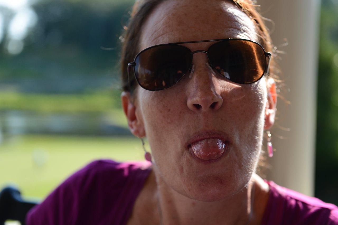 A person wearing sunglasses and a purple top sticks out their tongue playfully in an outdoor setting.
