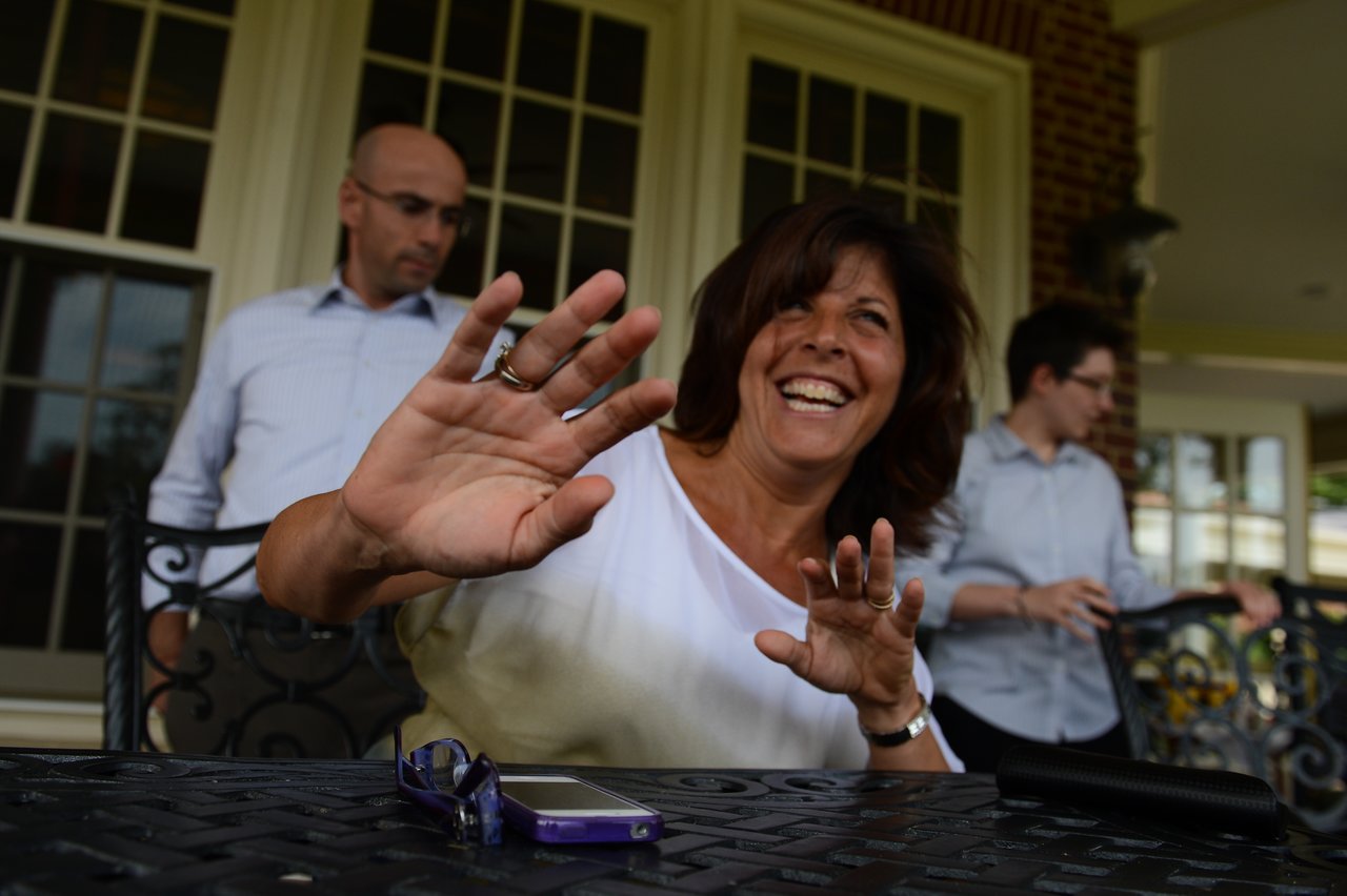 A smiling woman raises her hands playfully while sitting at a table, with two people standing behind her.
