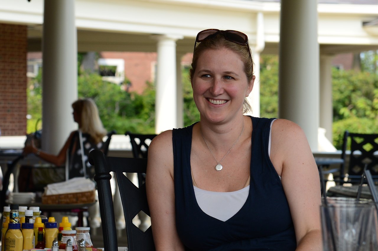 A woman with sunglasses on her head smiles while sitting at an outdoor table with drinks and condiments.