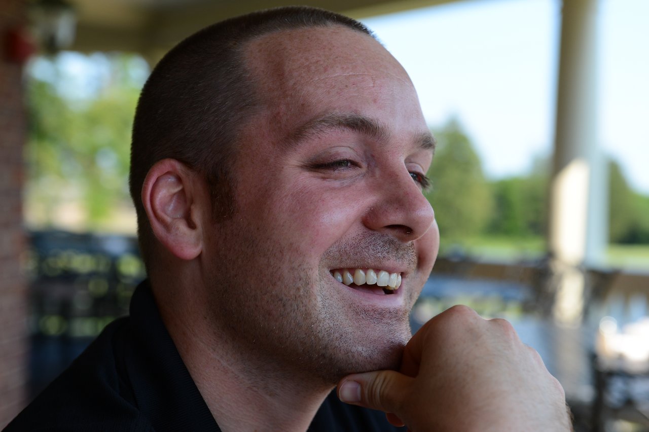 A man with a shaved head is smiling and resting his chin on his hand in an outdoor setting.