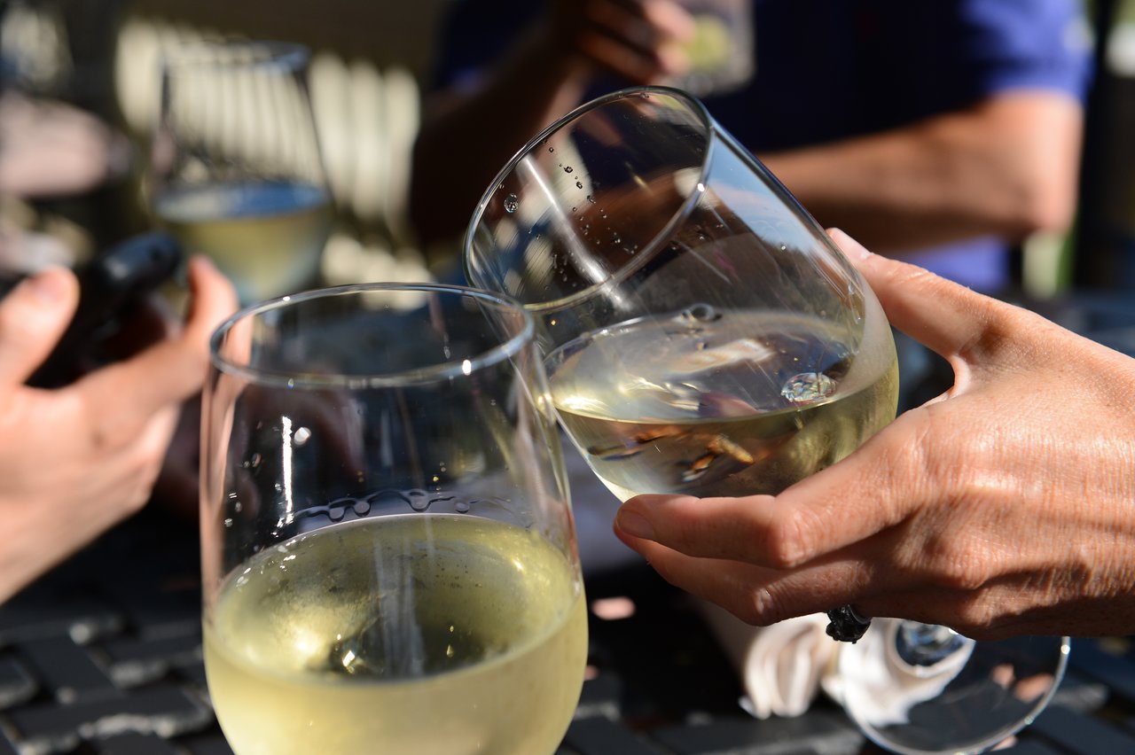 Two people clink glasses of white wine together while others hold drinks in the background.