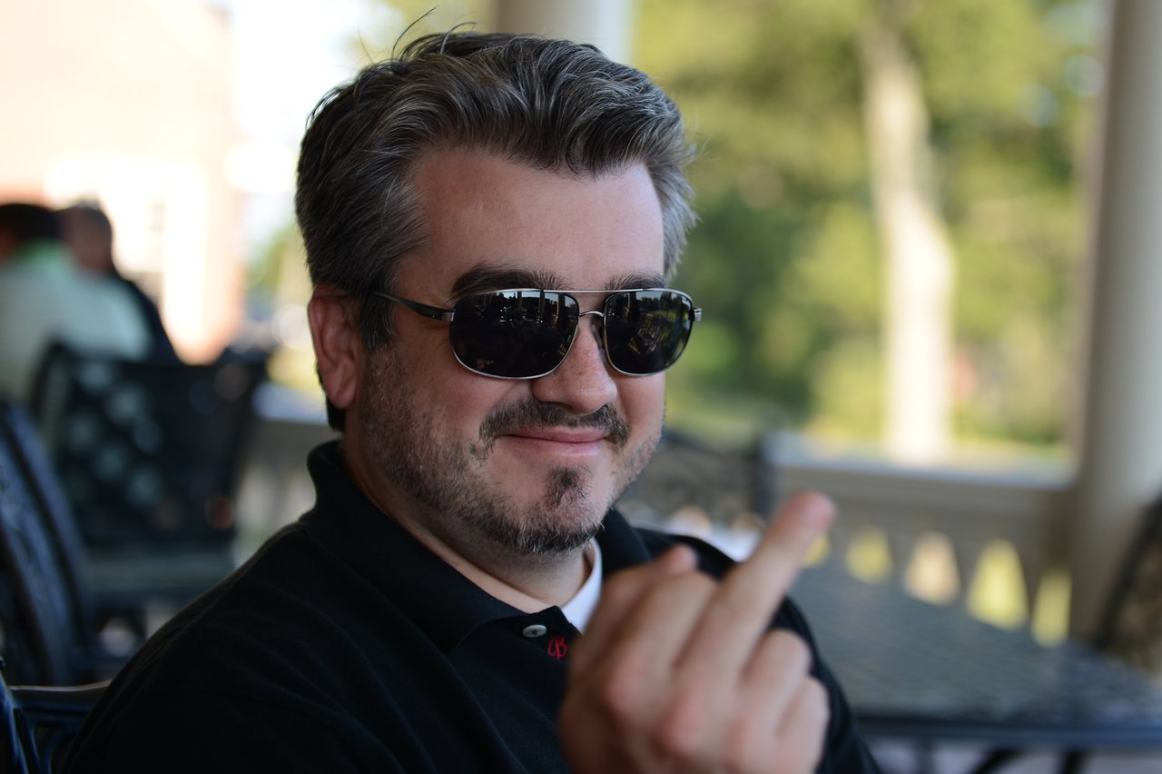 A man wearing sunglasses sits outdoors, smiling and making a rude hand gesture toward the camera.