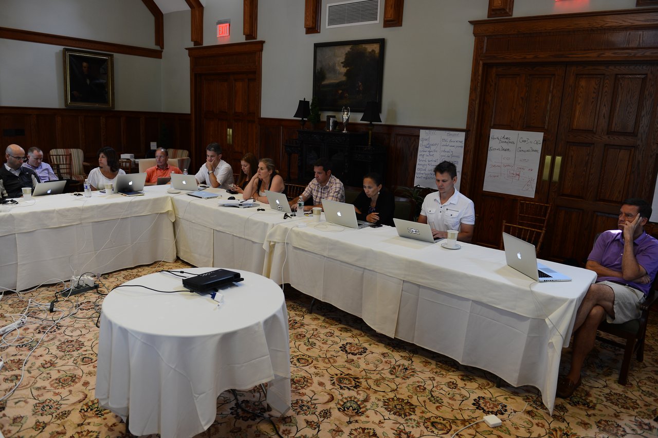 A group of people sit around tables with laptops, engaged in a meeting and discussion.