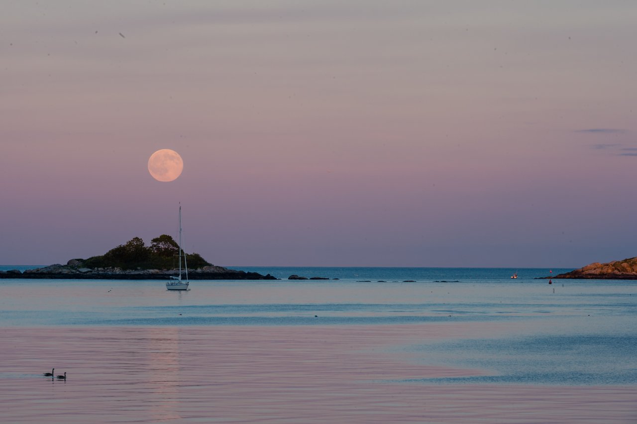 A full moon rises over the ocean near a small island, with a sailboat floating in the water.