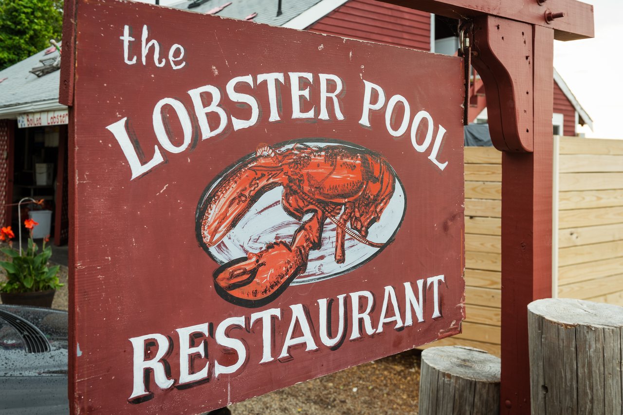 A wooden sign for "The Lobster Pool Restaurant" with a painted lobster, located outside a red building.