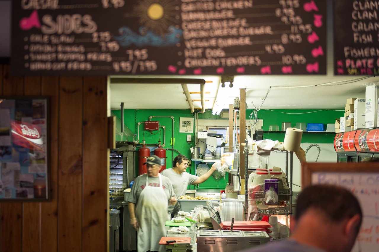 Two workers in a seafood restaurant kitchen prepare food, while a customer waits at the counter.