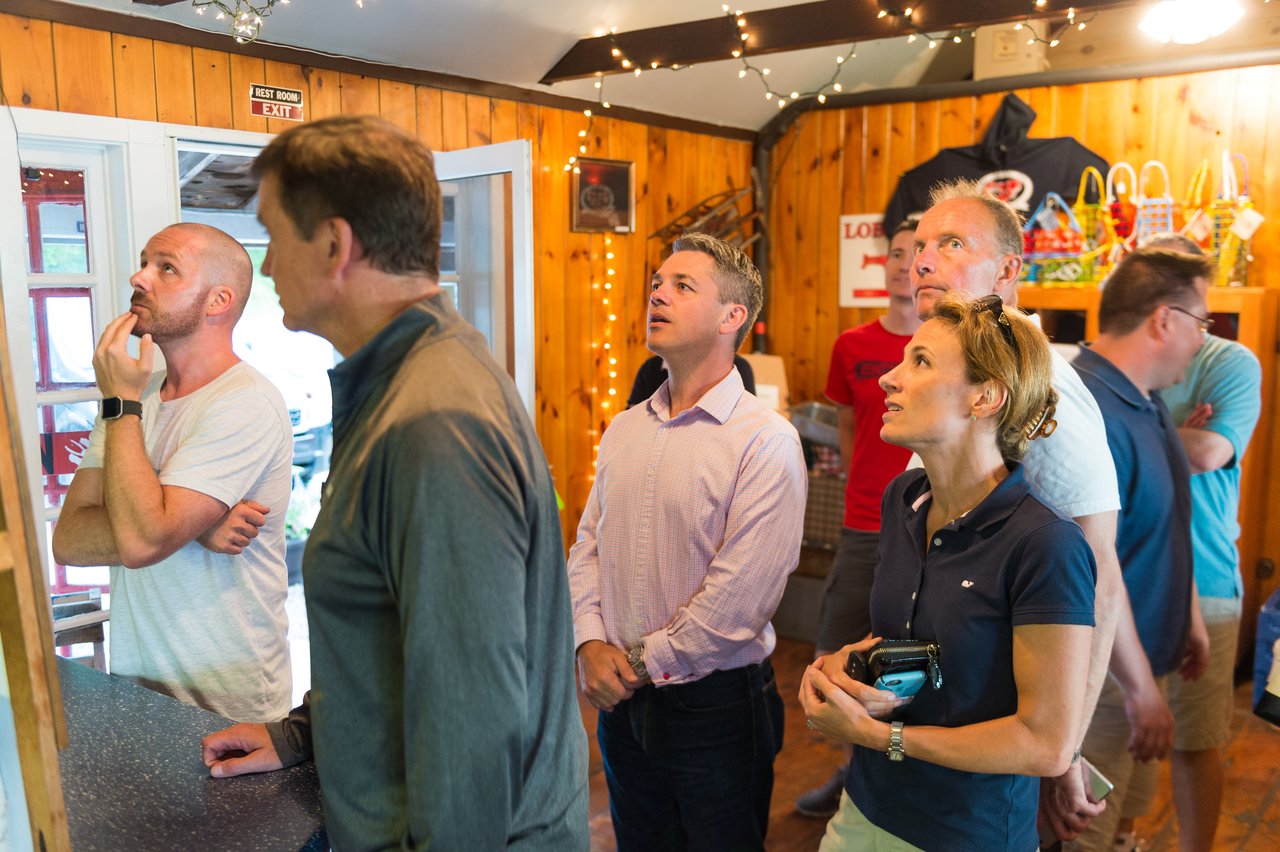 A group of people inside a wooden restaurant look up at a menu, appearing to decide what to order.