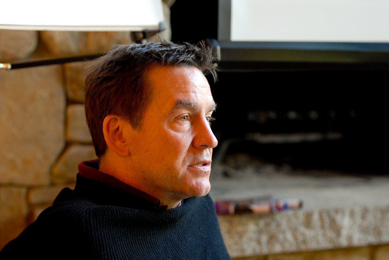 A man in a black sweater sits indoors, looking to the side with a focused expression.