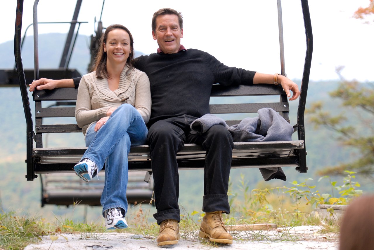 Two people sit on a ski lift, smiling and relaxed, with a scenic mountain view in the background.