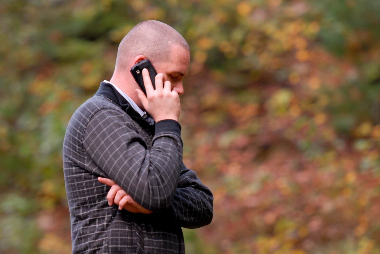 A person with a shaved head talks on a mobile phone while standing outdoors with arms crossed.