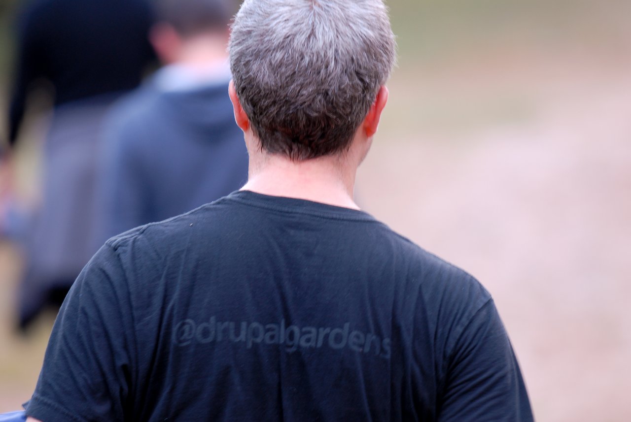 A person wearing a black "@drupalgardens" T-shirt walks outdoors with others in the background.